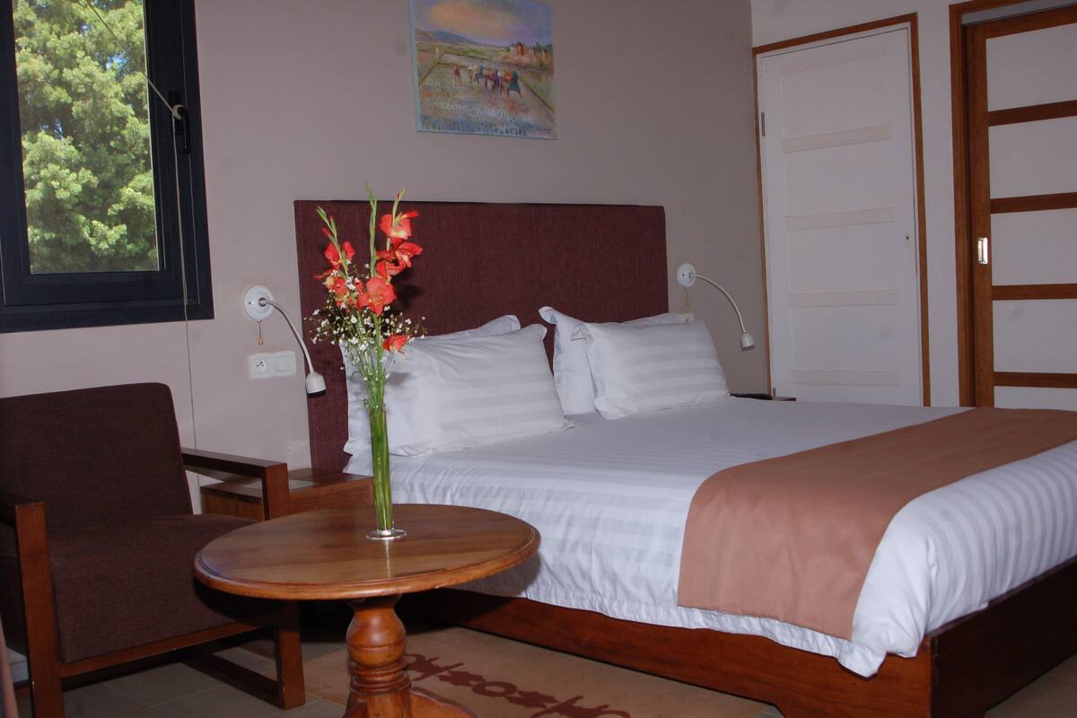 A hotel room with a bed, a chair, a round wooden table, and a vase with flowers.