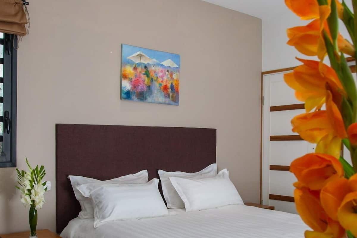 A bedroom with a wooden bed, white bedding, a small bedside table with a vase of flowers, and a painting on the wall.