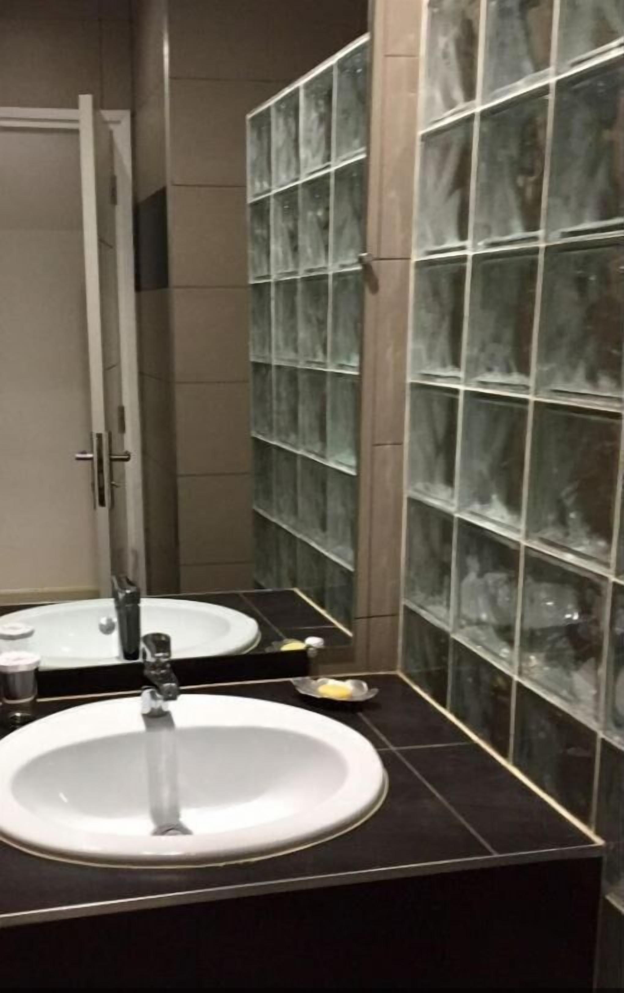 Un baño con un lavamanos blanco, un espejo y una pared de bloques de vidrio.