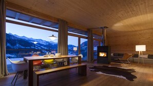Un interior moderno con techo de madera, ventanas amplias con vistas a la montaña, zona de comedor con mesa y sillas, y una chimenea.