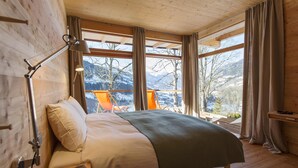 Un dormitorio de cabaña de madera con una cama, una mesita de noche, una lámpara de lectura y vistas a montañas nevadas a través de grandes ventanales.