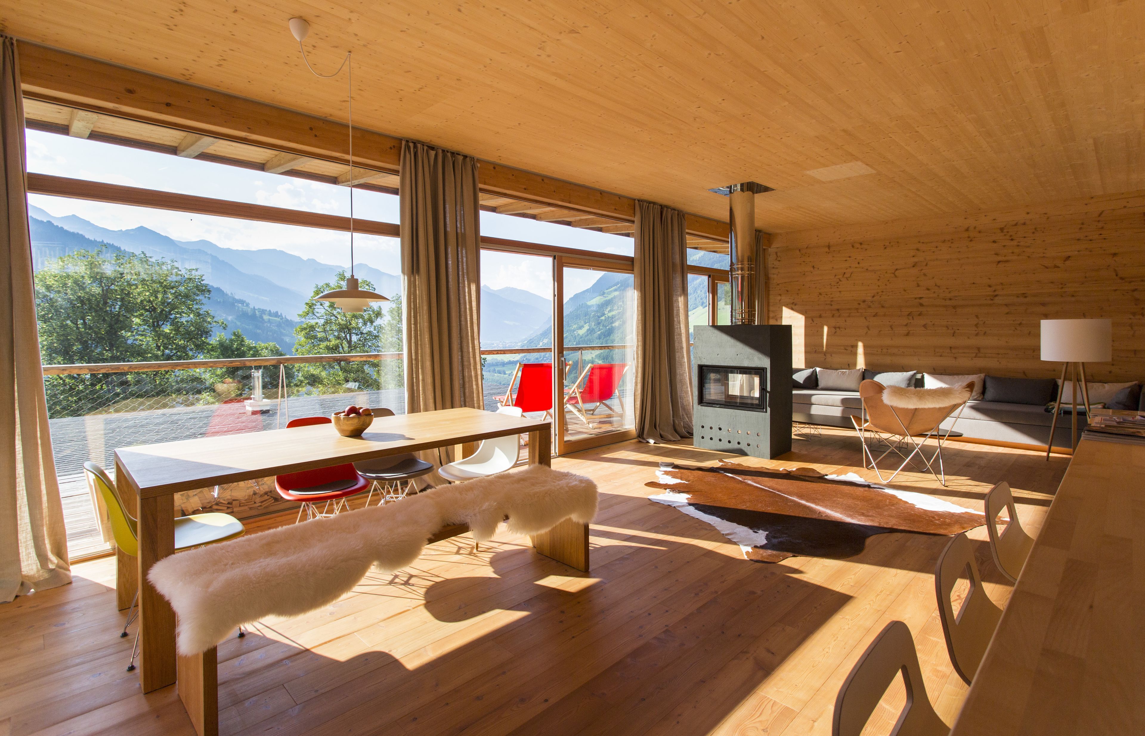 Un interior moderno con suelo de madera, amplias ventanas que ofrecen vistas a las montañas, una zona de comedor con sillas rojas y un rincón de descanso acogedor con chimenea.