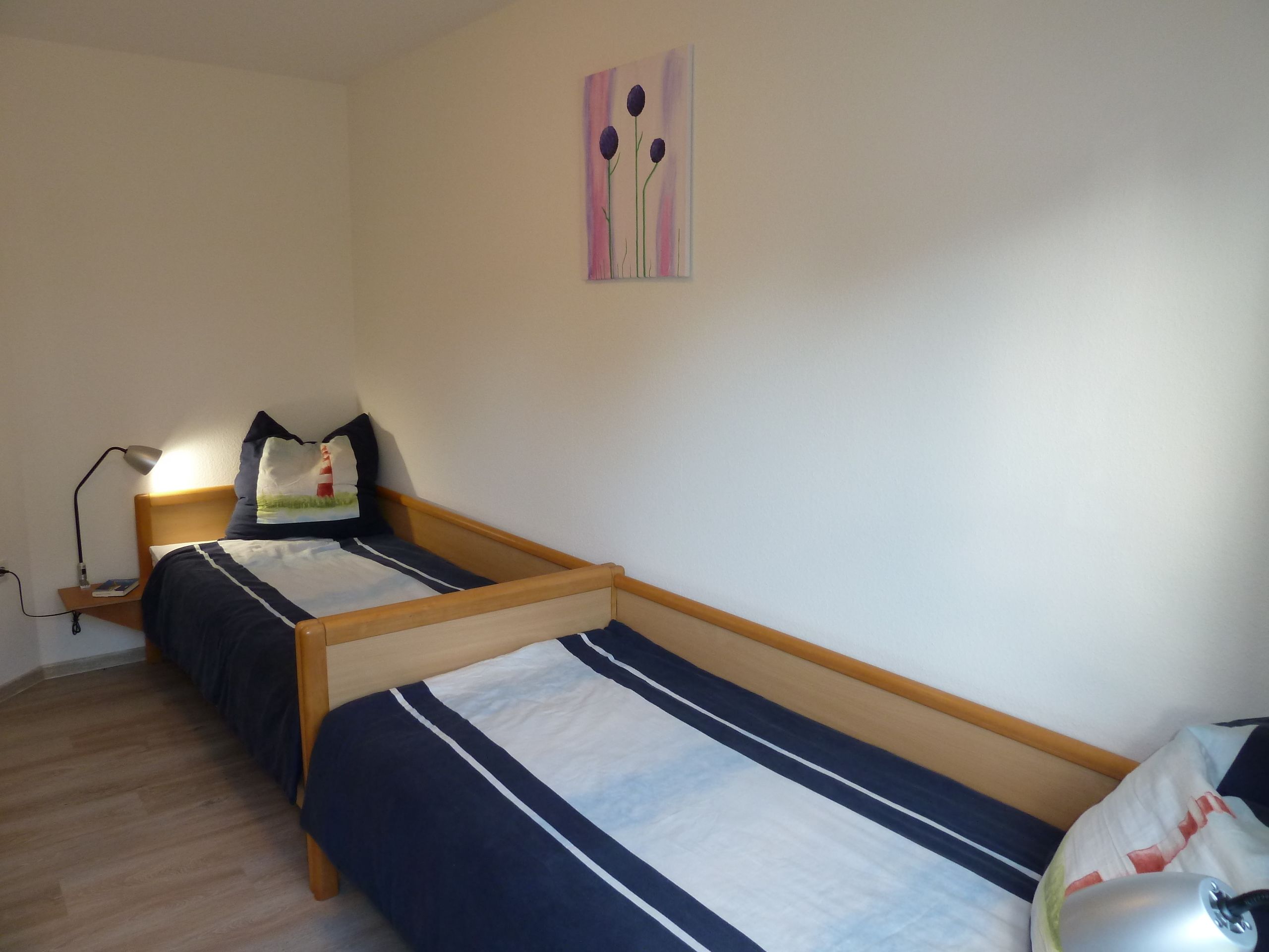 A small, clean room with two single beds, a bedside table with a lamp, and a wall-mounted painting.