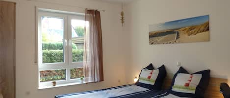 Un dormitorio con cama, ventana con cortinas, lámpara de pared y una pintura de una escena de playa.
