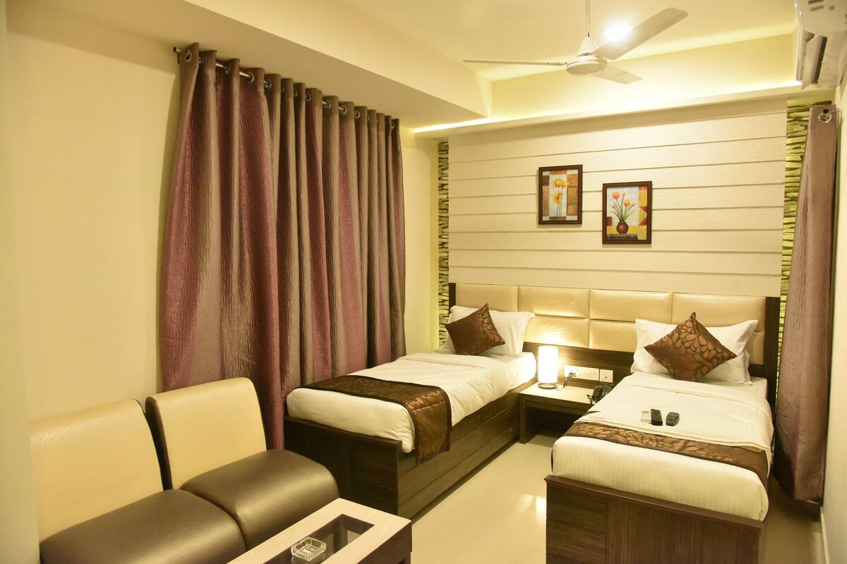A hotel room with two beds, a sofa, a bedside table with a lamp, and a ceiling fan.