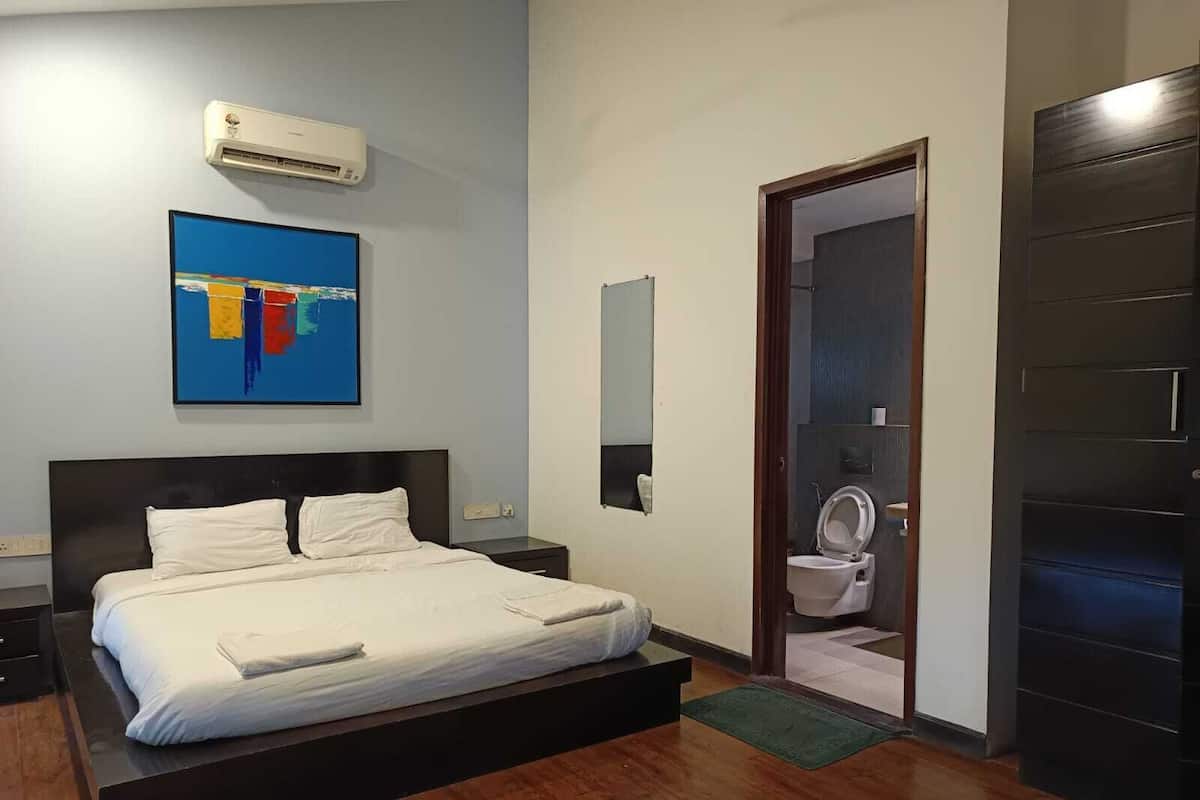 A modern bedroom with a large bed, a nightstand, a wall-mounted air conditioner, a colorful abstract painting, a mirror, and a door leading to a bathroom.