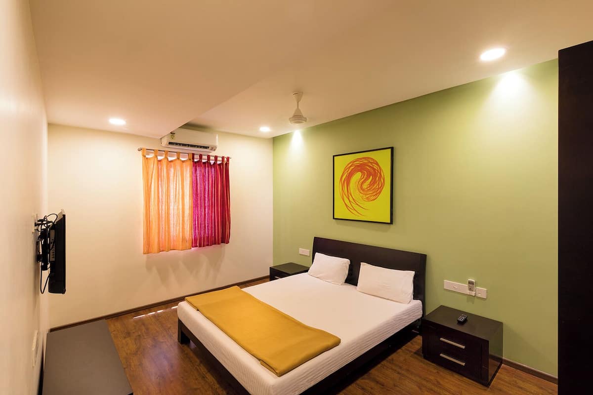 A hotel room with a bed, a nightstand, a wall-mounted air conditioner, a window with colorful curtains, and a painting on the wall.