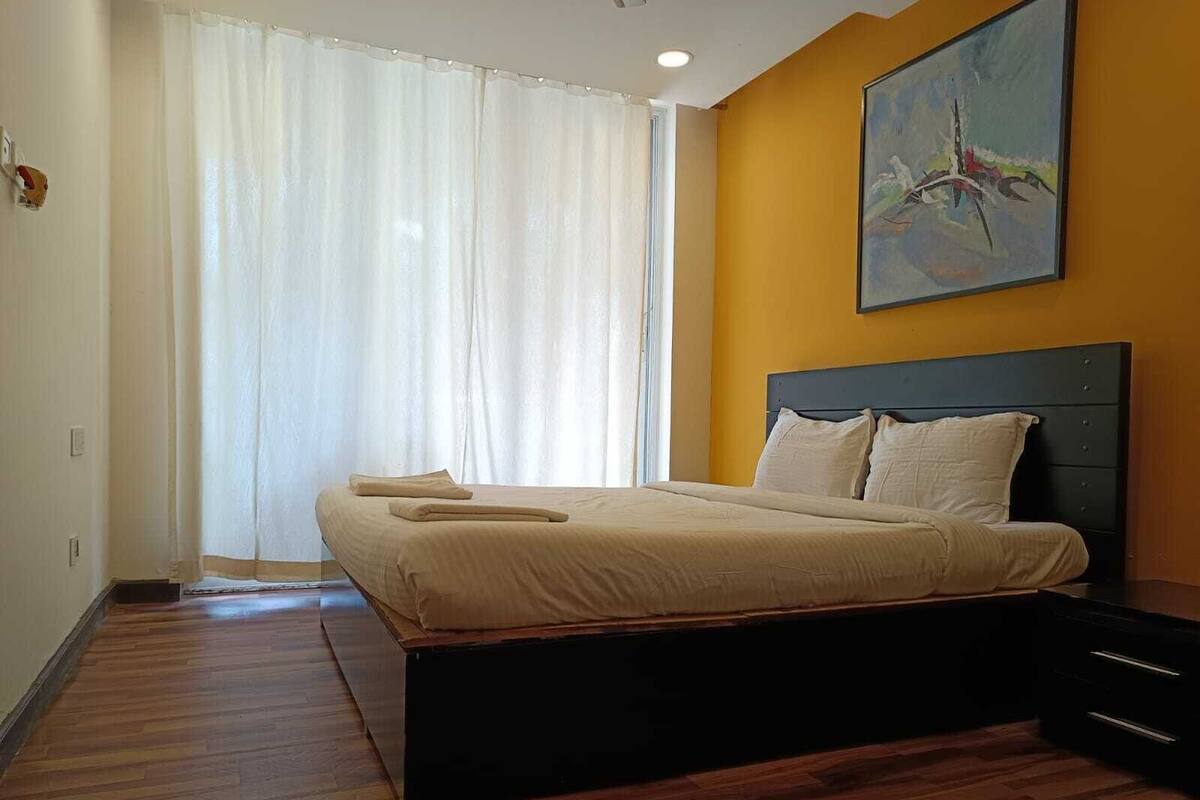 A bedroom with a bed, a nightstand, a painting, and a window with curtains.