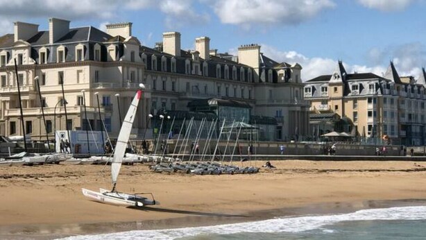 A beach with a sailboat, a sandy shore, and buildings in the background.