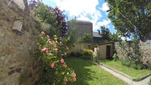 A stone wall with a birdhouse, a small garden with pink flowers, and a building with a door and window.