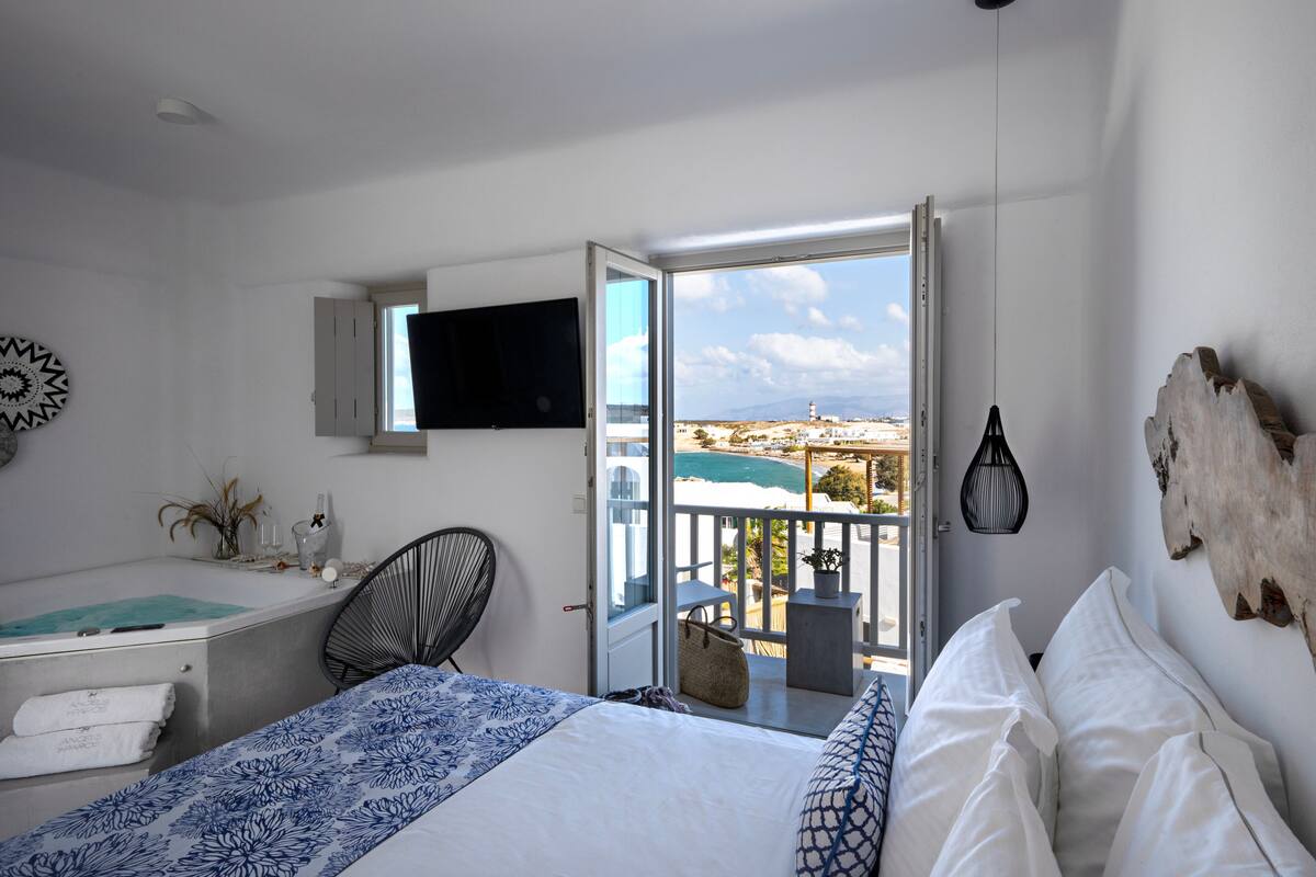 A bedroom with a bed, a desk, a chair, a TV, and a balcony with a view of the sea.