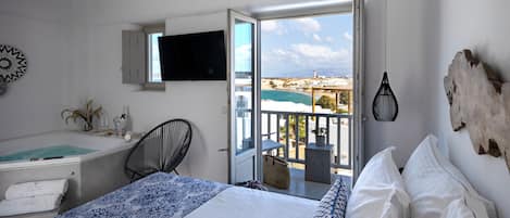 A bedroom with a bed, a desk, a chair, a TV, and a balcony with a view of the sea.
