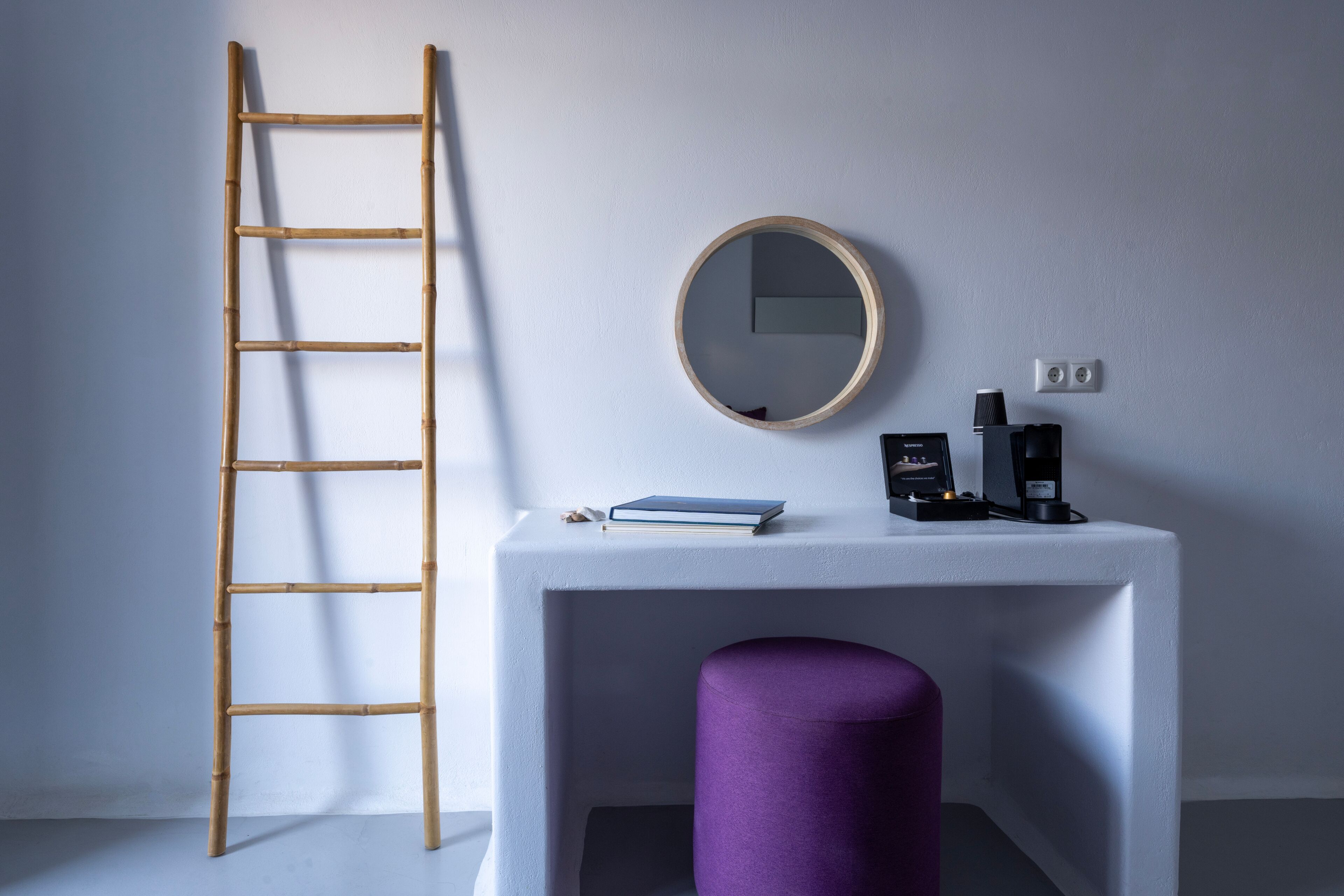 Een kamer met een wit bureau, een paarse kruk, een ronde spiegel, een ladder en een koffieapparaat.