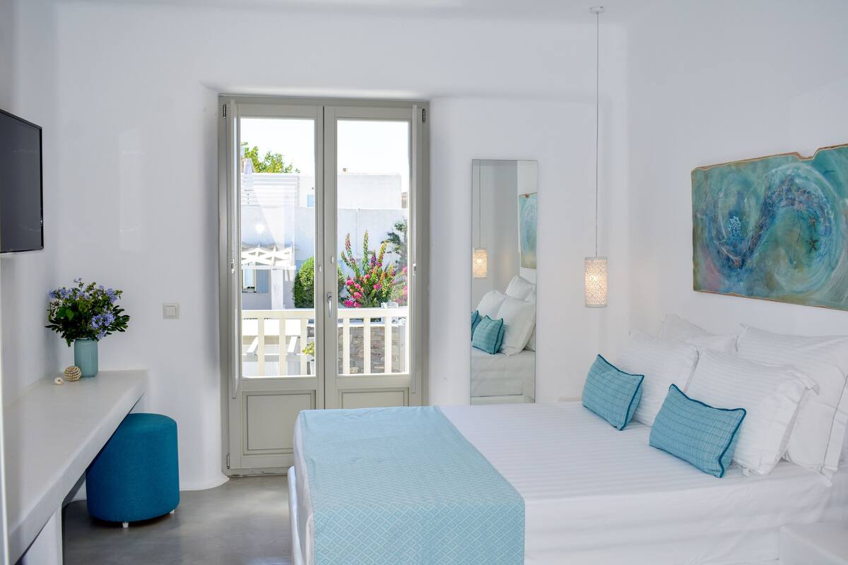 A modern bedroom with a large bed, a blue ottoman, a TV, and a balcony with flowers.