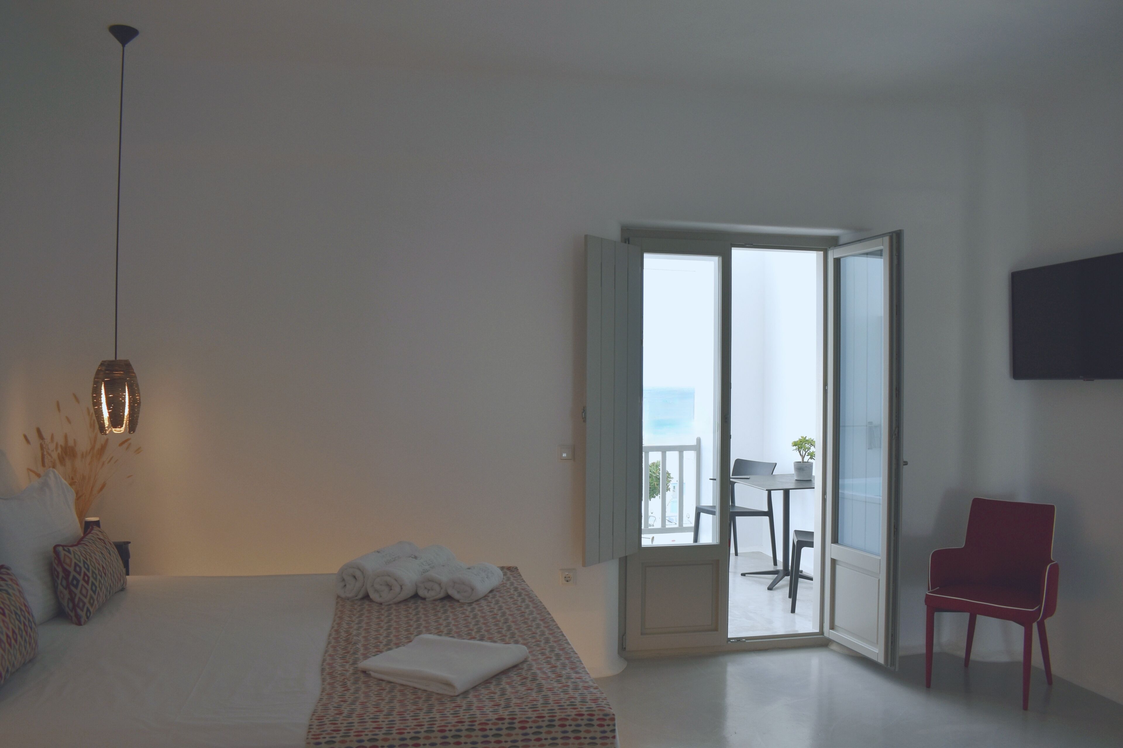Habitación de hotel con cama, una silla roja, una mesa con planta y un balcón con vistas.