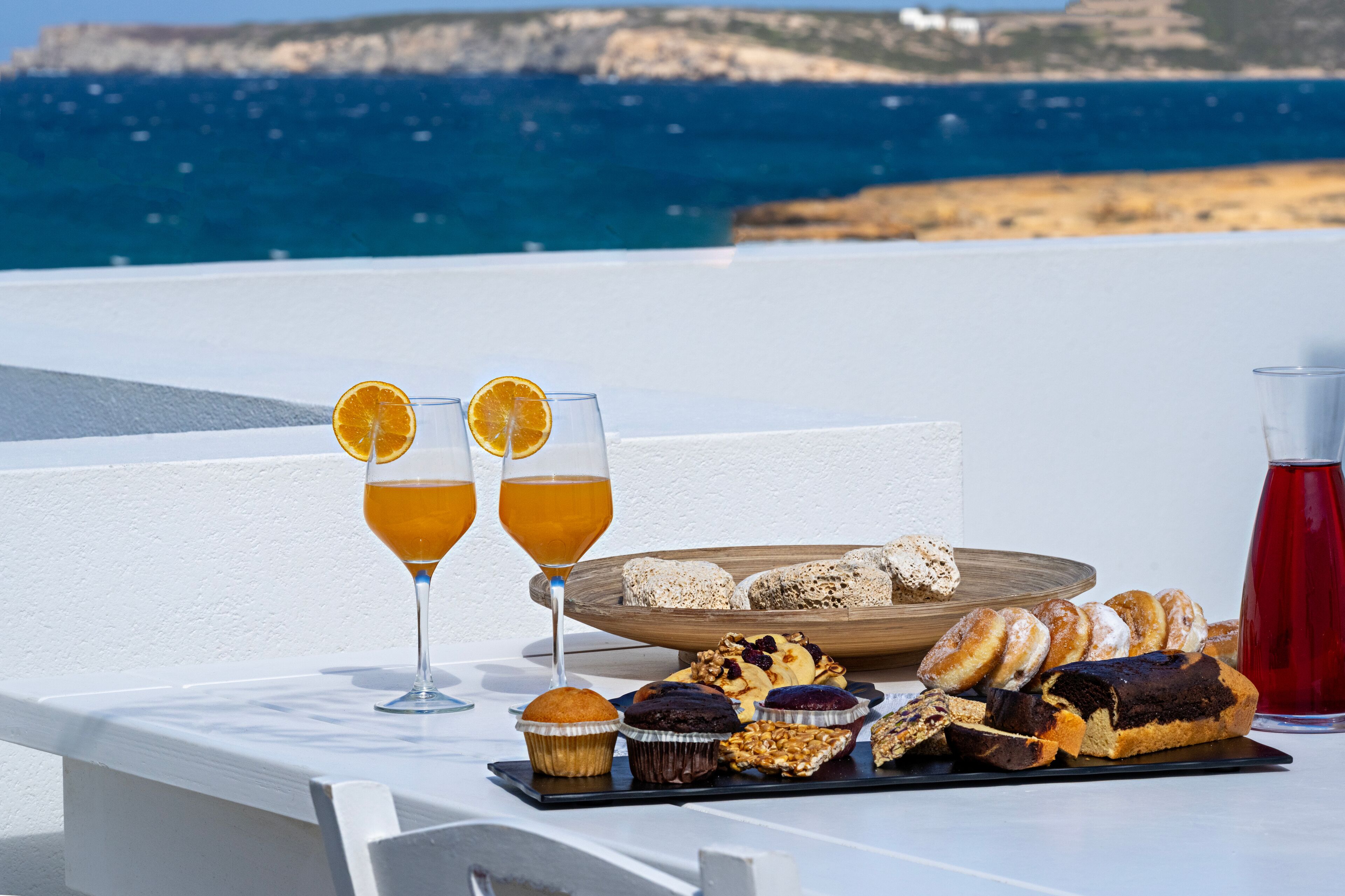 A table with two glasses of orange juice, a tray of pastries, and a red drink, set against a coastal backdrop.
