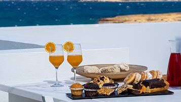 A table with two glasses of orange juice, a tray of pastries, and a red drink, set against a coastal backdrop.