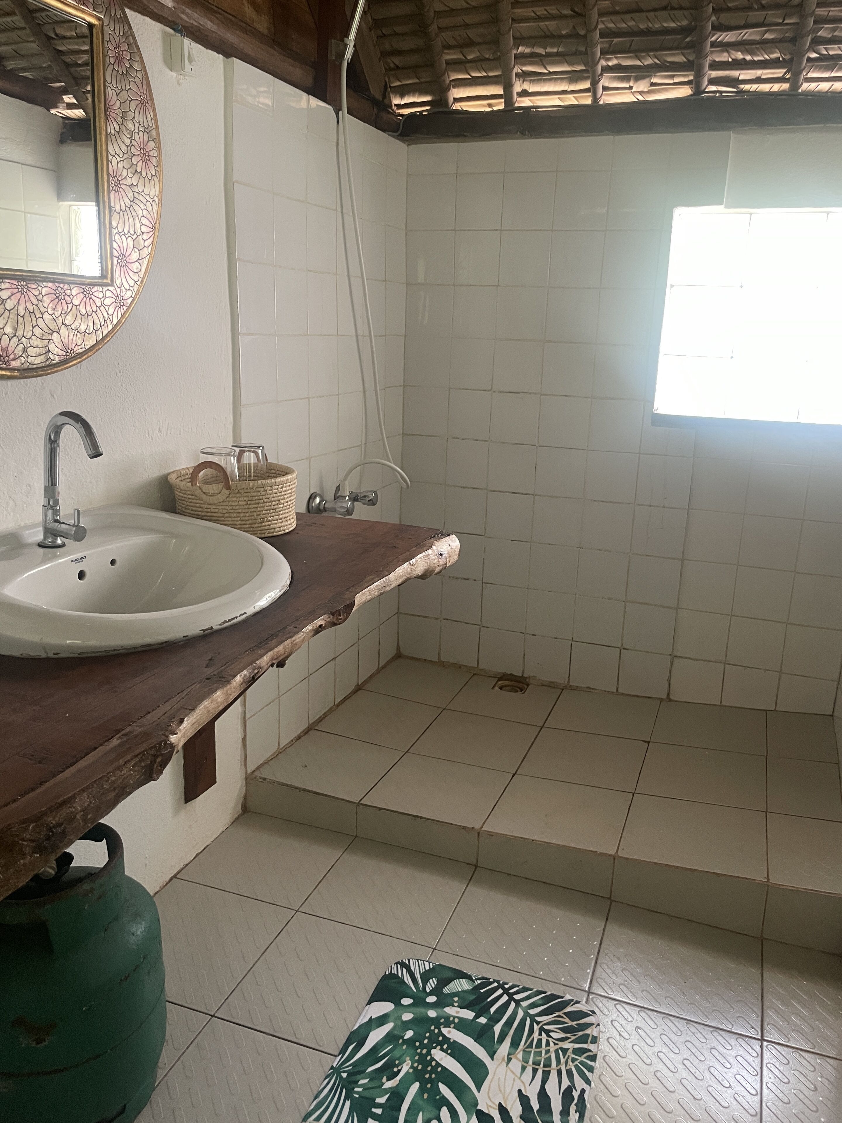 Un baño con lavamanos blanco, espejo con marco floral, ventana y suelo de baldosas.