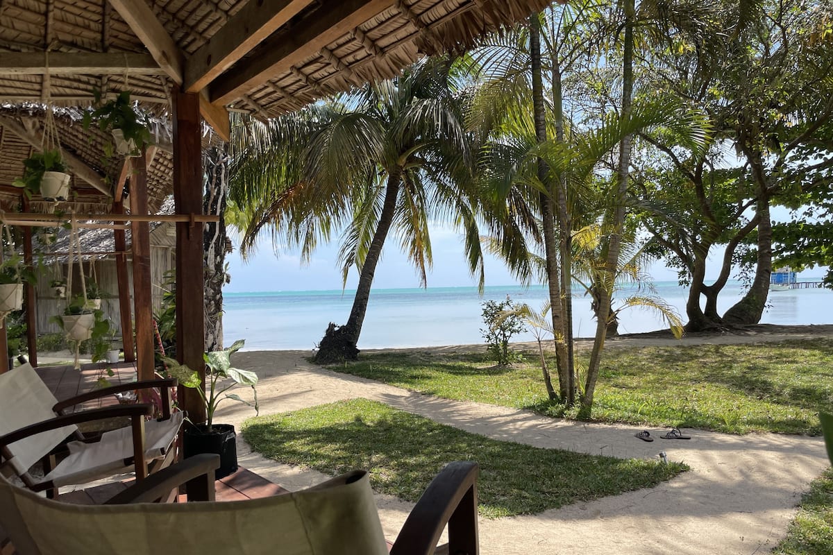 Eine Terrasse am Strand mit einem Reetdach, Palmen und Blick auf das Meer.