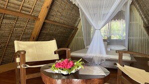 Una habitación con techo de paja, muebles de madera, una cama con dosel blanco y una mesa con un ramo de flores.