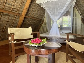 A room with a thatched roof, wooden furniture, a bed with a white canopy, and a table with a bouquet of flowers.