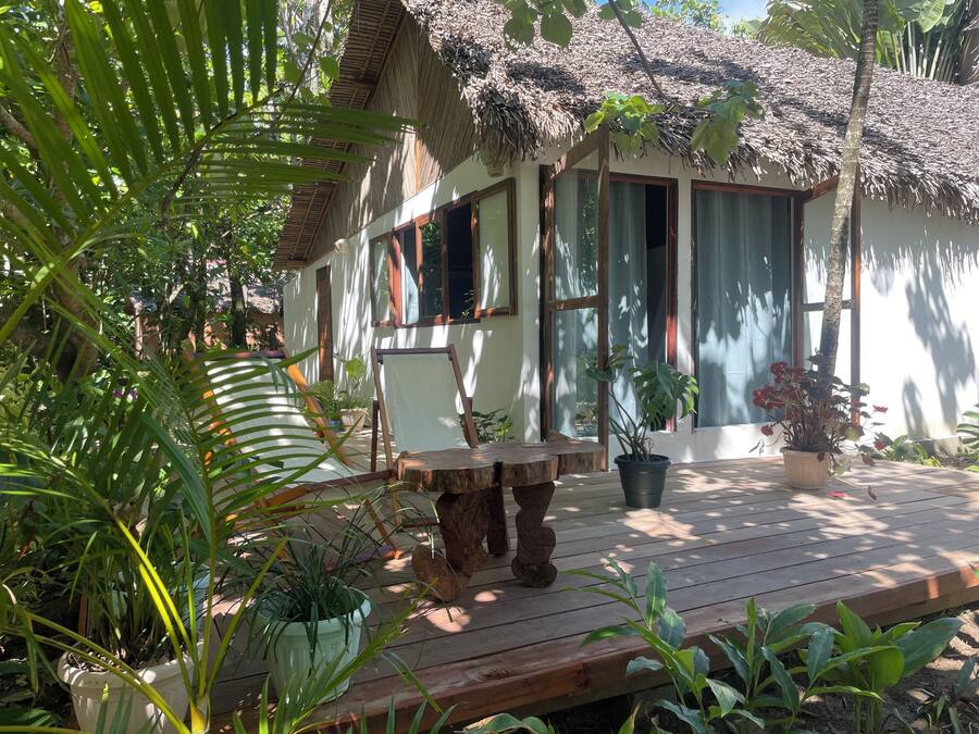 A wooden deck with a table and chairs, surrounded by potted plants and a thatched-roof building.