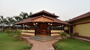 A covered outdoor seating area with a wooden structure and a brick pathway leading to it. - The Kanila Resort (Karjat)
