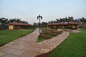 A pathway leading to a row of yellow buildings with red roofs, surrounded by well-maintained grass and palm trees. - The Kanila Resort (Karjat)