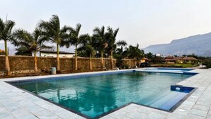 A rectangular swimming pool with clear blue water, surrounded by a tiled deck. Palm trees are visible in the background, and there are buildings with red roofs in the distance. - The Kanila Resort (Karjat)