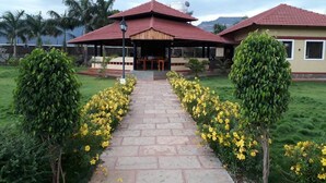 A pathway leading to a building with a red roof, surrounded by greenery and yellow flowers. - The Kanila Resort (Karjat)