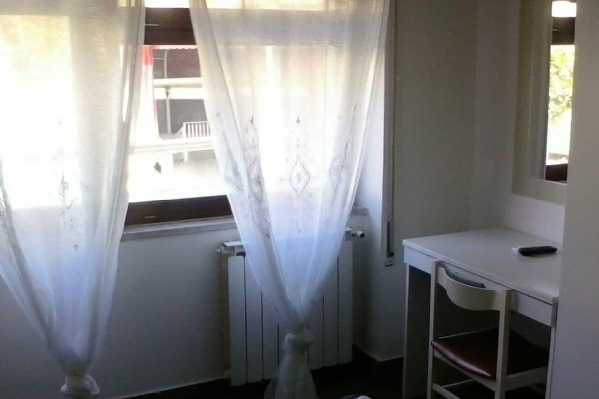 A small, compact room with a single bed, a bedside table, and a window with sheer curtains.