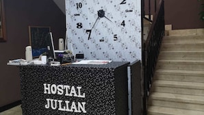 A reception desk labeled "HOSTAL JULIAN" with a clock above it, a framed picture on the wall, and a staircase to the right.