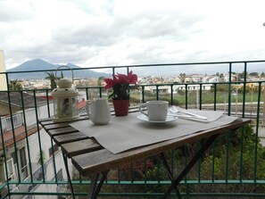 Uma varanda com mesa posta para dois, um vaso de planta e vista para a cidade e montanhas.