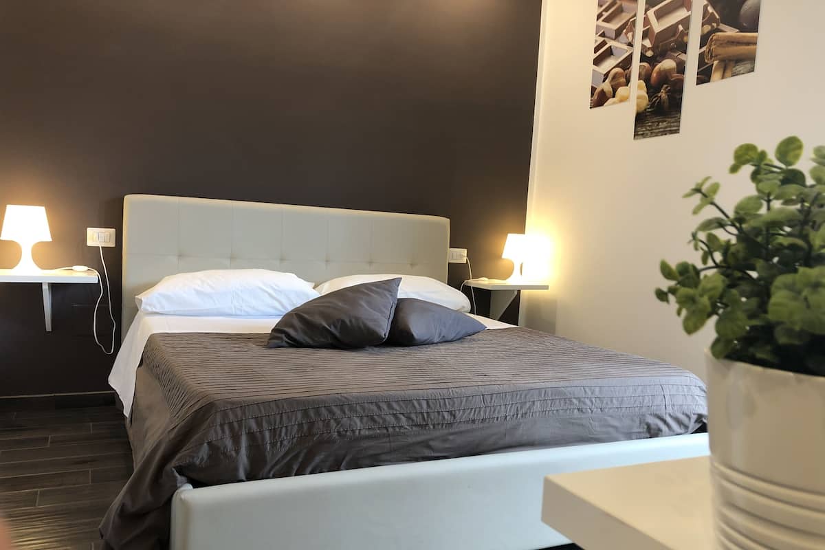 A modern bedroom with a bed, bedside lamps, a nightstand, and a potted plant.
