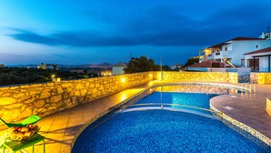A swimming pool with stone walls and a view of the city at dusk. - Villa Alysso (Chania)