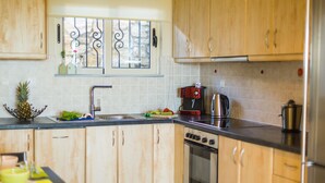 A kitchen with wooden cabinets, a stainless steel refrigerator, a stove, and a window with decorative iron bars. - Villa Alysso (Chania)