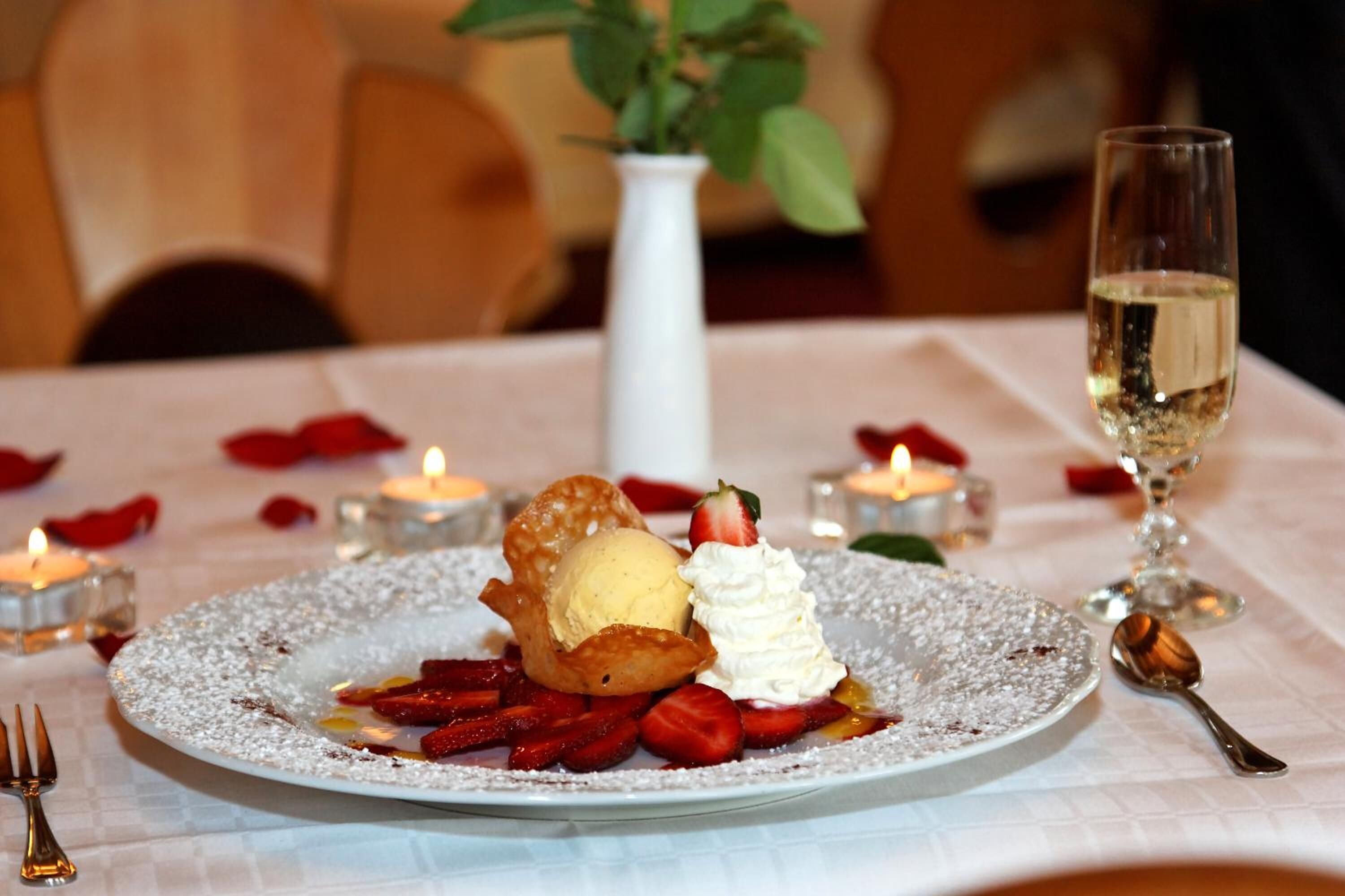 A dessert plate with strawberries, whipped cream, and a scoop of ice cream, accompanied by a glass of white wine and candles on a table.