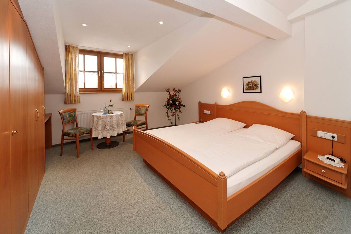 A bedroom with a large bed, a wooden wardrobe, a small table with chairs, and a window with curtains.