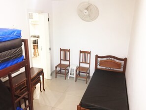 Habitació petita i senzilla, amb un llit individual, dues cadires de fusta, un escriptori i un ventilador.