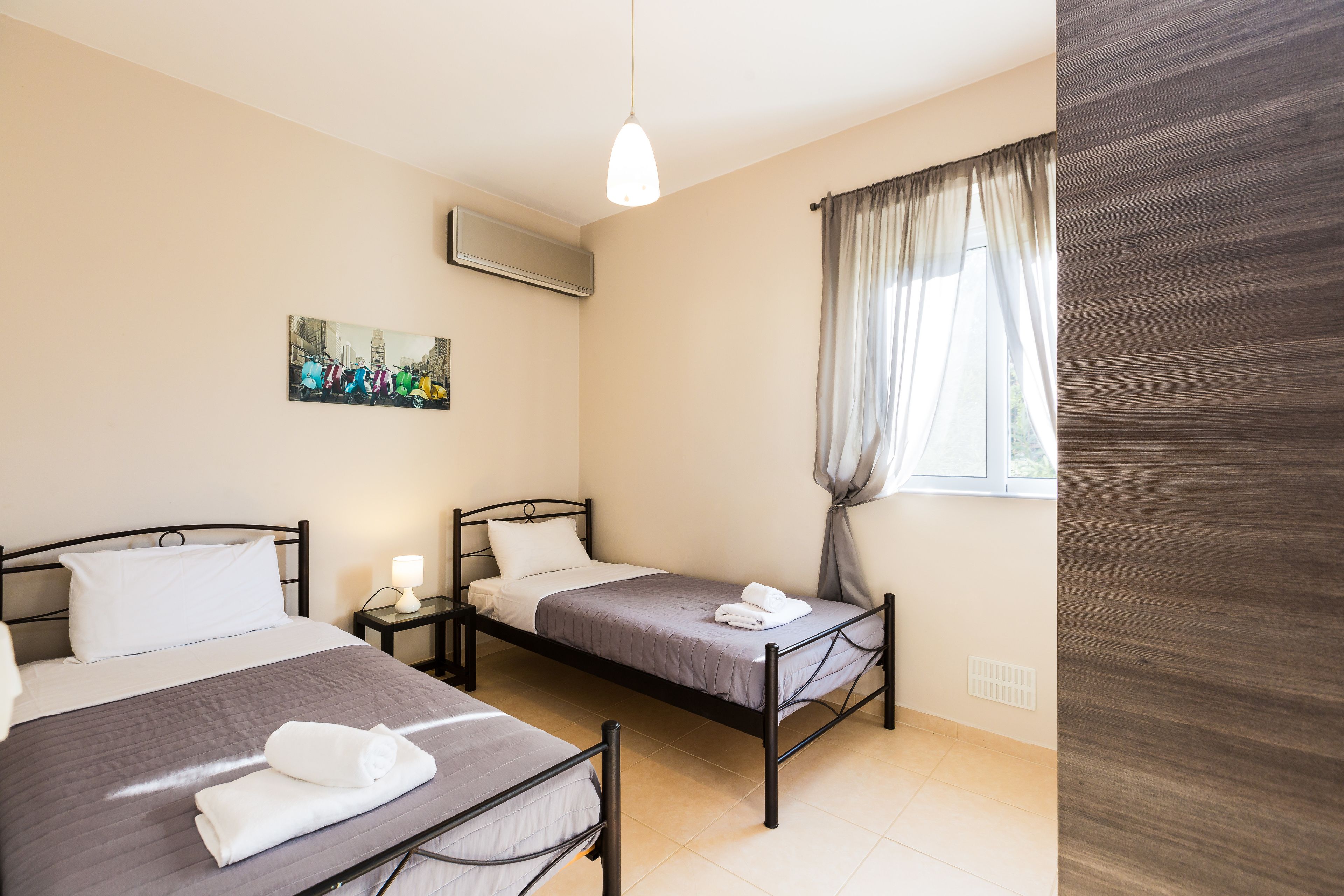 Een hotelkamer met twee enkele bedden, een nachtkastje met een lamp, een schilderij aan de muur en een raam met gordijnen.
