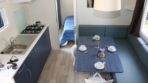 A compact kitchenette with a stove and sink, a dining table set for four with cups and saucers, and a bench with cushions. - Happy Camp Mobile Homes In Karda Beach Camping and Bungalows (Corfu)