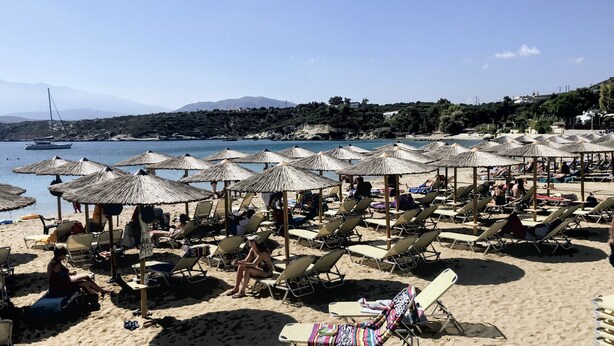 Ein Strand mit Reihen von Liegestühlen und Reetsonnen schirmen, ein Boot im Wasser und eine bergige Kulisse.