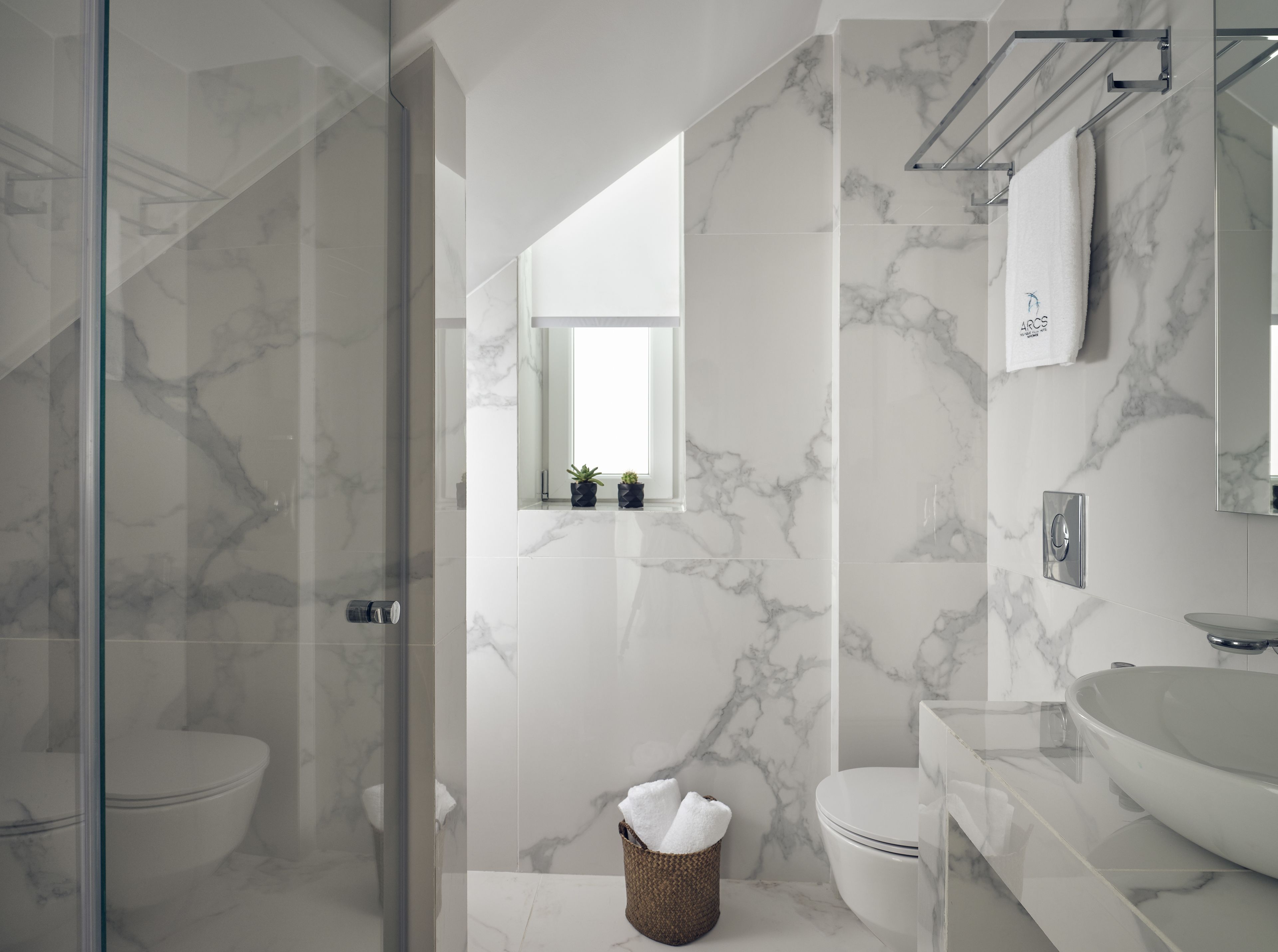 Ein modernes Badezimmer mit Duschkabine aus Glas, einer weißen Toilette, einer Wand mit Marmorfliesen, einem Waschbecken und einem kleinen Fenster mit Pflanzen.