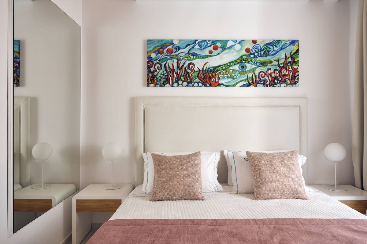 Un dormitorio moderno con una cama grande, dos mesitas de noche, una mesilla auxiliar, un cuadro abstracto de gran tamaño y un espejo.