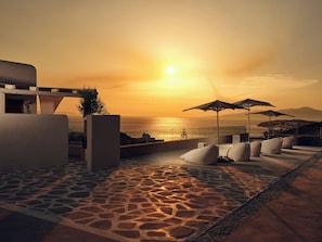 A rooftop terrace with a stone floor, lounge chairs, and umbrellas overlooking the sea at sunset. - ΑRCS Suites & Residences (Mykonos)