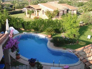 A swimming pool surrounded by a garden with various plants and a house in the background. - Villa con Piscina Miriacheddu (San Teodoro)