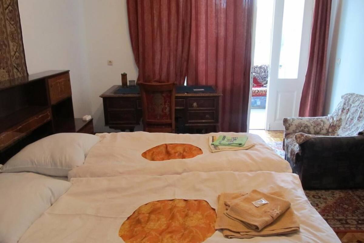 A hotel room with two beds, a wooden dresser, a chair, and a view of the outside through a window with red curtains.