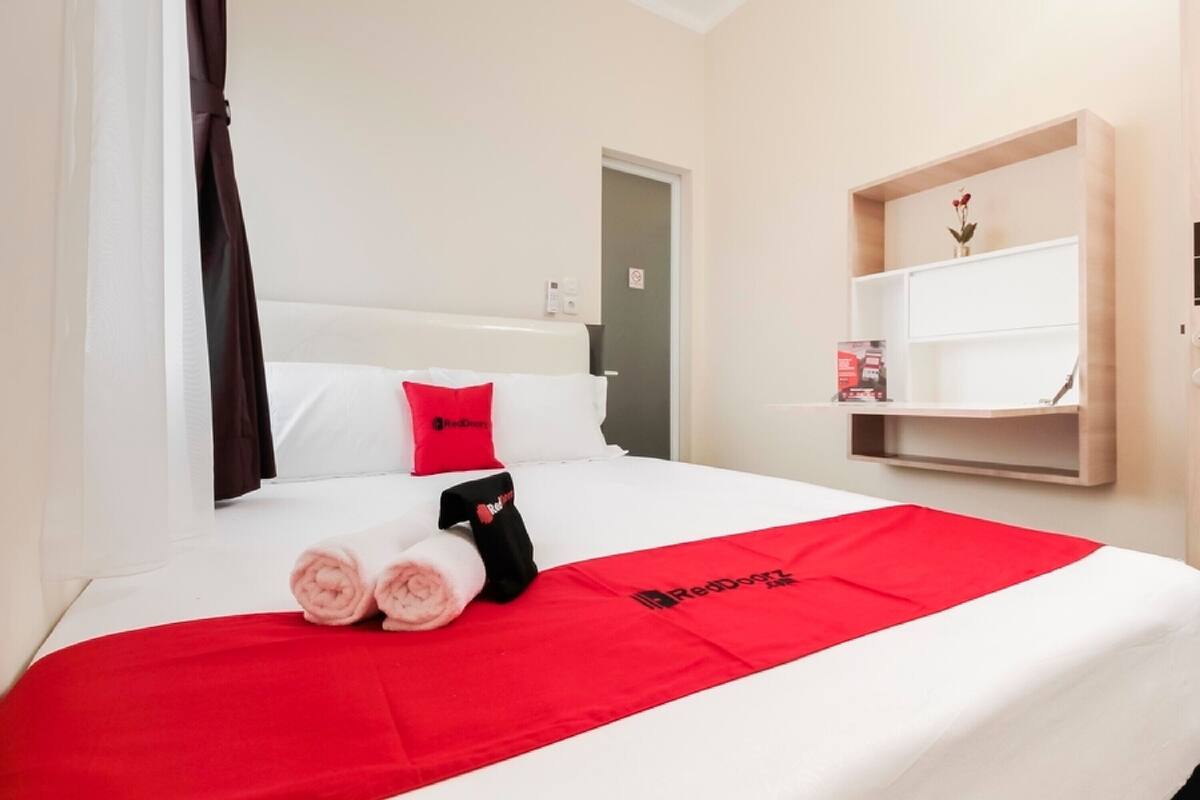 A hotel room with a bed, a red and white bedspread, a red pillow, a black towel, and a shelf with a vase.