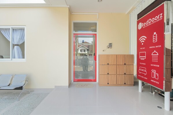 A RedDoorz hotel entrance with a glass door, a waiting area with a bench, and a sign listing amenities like free WiFi, mineral water, television, and bathroom. - RedDoorz near Soekarno Hatta Airport (Tangerang)
