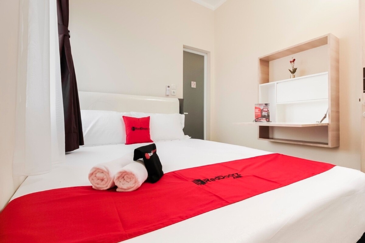 A hotel room with a bed, a red and white bedspread, a red pillow, a black towel, and a shelf with a vase.